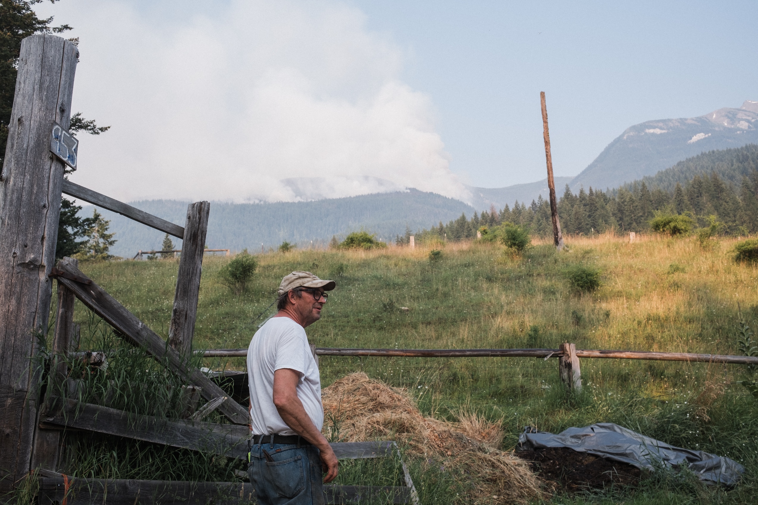 A man stands next to a fence in a grass meadow. Wildfire smoke rises from hills in the distance