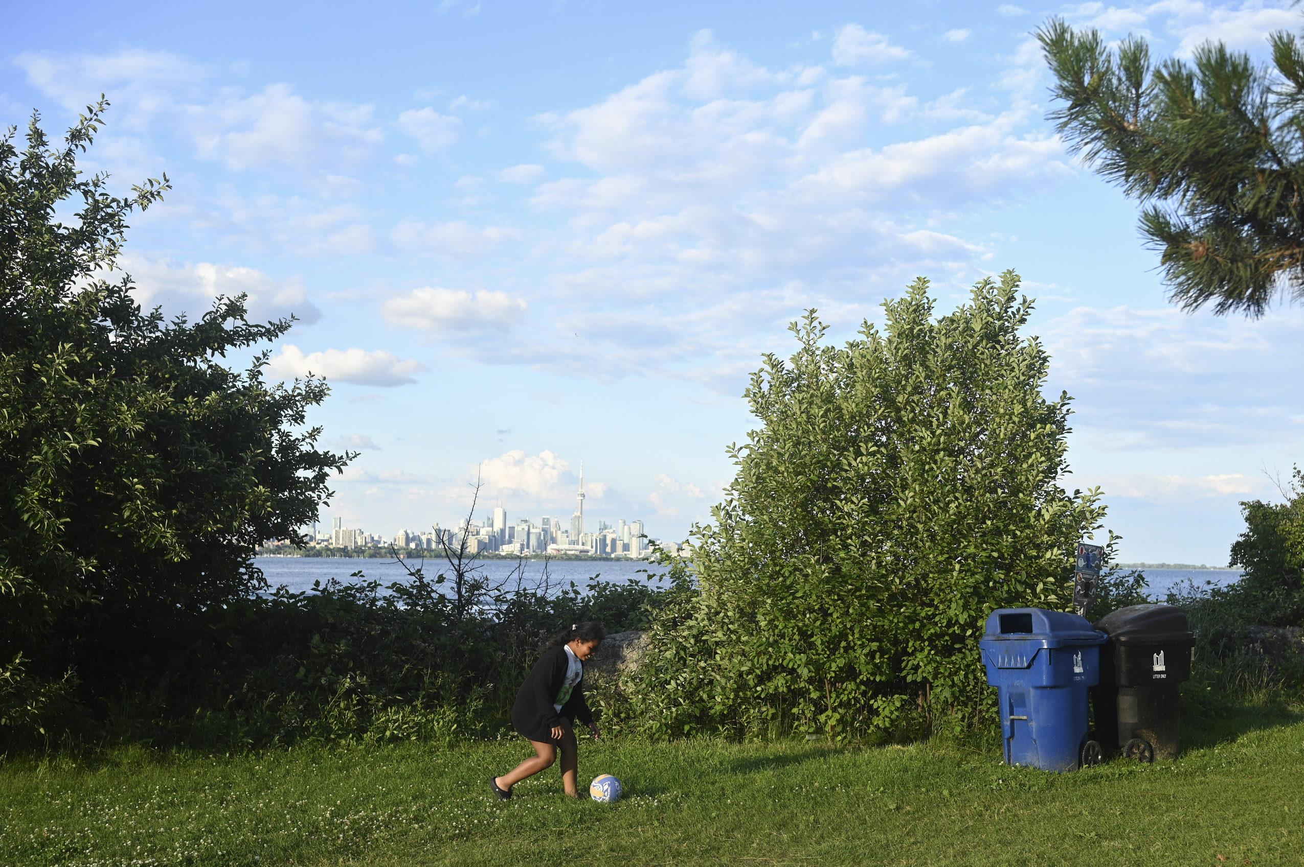 A child plays soccer on grass at Humber Bay Park with Lake Ontario and the downtown Toronto skyline visible in the background
