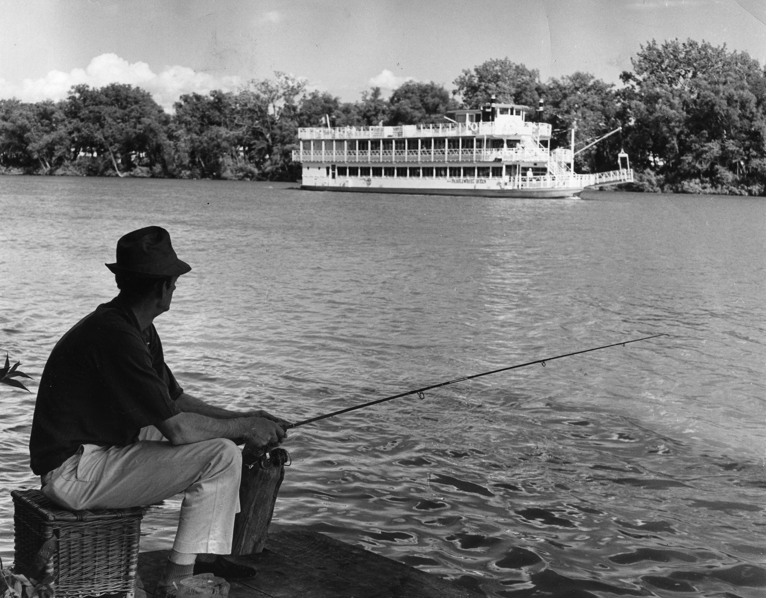 Black and white photo of a man fishing near a river with a paddlewheeler visible